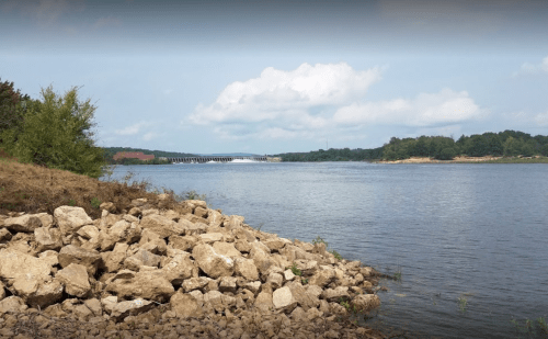 A calm river scene with rocky shorelines, green trees, and a bridge in the distance under a partly cloudy sky.