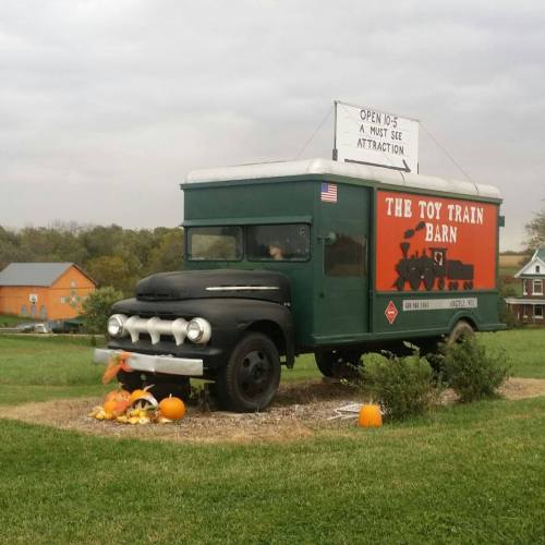 A vintage truck converted into a toy train barn, surrounded by pumpkins and grass under a cloudy sky.