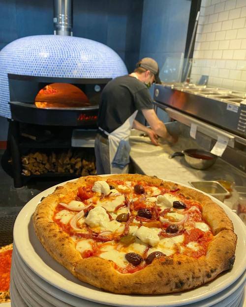 A freshly baked pizza with toppings sits on a plate in front of a chef working in a restaurant kitchen.