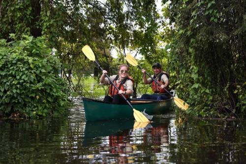 Two people in a green kayak paddling through a lush, green waterway surrounded by trees and foliage.