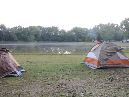 Two tents sit on grassy ground by a calm lake, with mist rising and trees in the background.