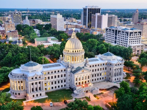 Aerial view of a grand government building with a dome, surrounded by city skyline and greenery at dusk.