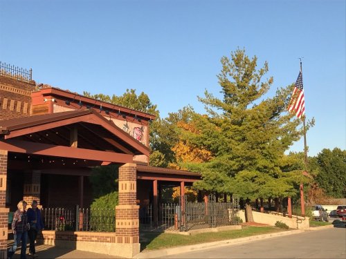 A brick building with a wooden awning, surrounded by trees and an American flag waving in the background.