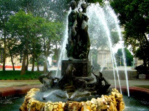 A decorative fountain featuring a statue of two figures surrounded by water jets and lush greenery.