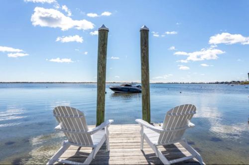 Two white Adirondack chairs on a dock overlooking a calm waterway with a boat in the distance under a blue sky.