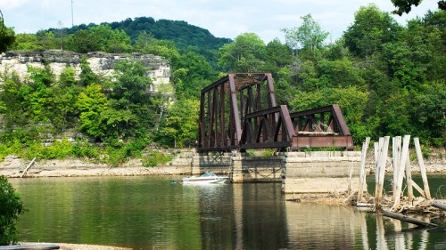 A rusted train bridge spans a calm river, surrounded by lush green trees and rocky cliffs in the background.