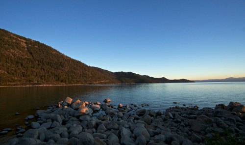 A serene lakeside view at sunset, featuring calm waters, rocky shore, and forested hills in the background.