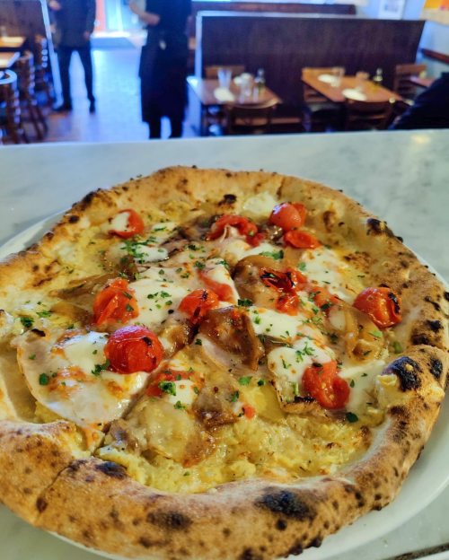 A close-up of a pizza topped with melted cheese, roasted tomatoes, and herbs, served on a marble countertop.