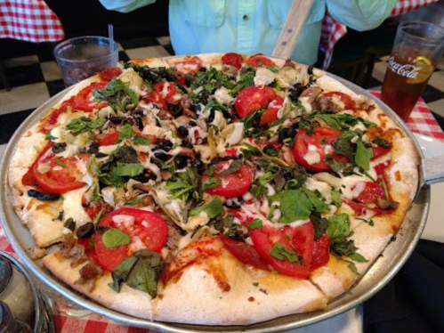 A large pizza topped with tomatoes, olives, greens, and various meats, served on a silver platter.