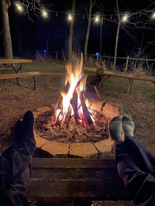 Two pairs of feet relax by a crackling campfire, surrounded by a dark outdoor setting with string lights in the background.