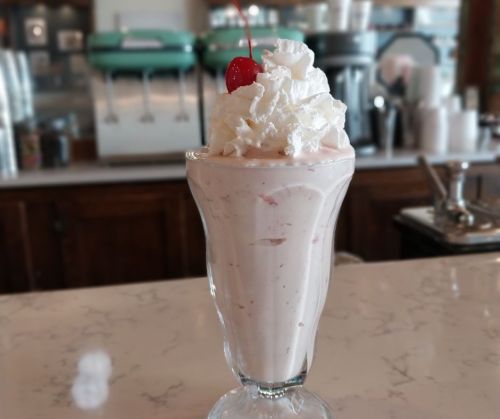 A tall glass of strawberry milkshake topped with whipped cream and a cherry, set on a marble countertop.