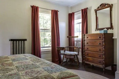 Cozy bedroom featuring a quilted bed, wooden dresser, rocking chair, and large windows with red curtains.