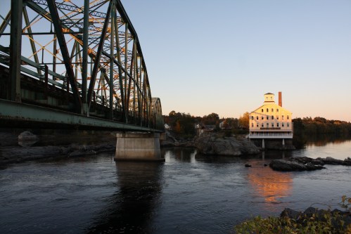 A green metal bridge spans a river, with a white building on a rocky shore reflecting the sunset.