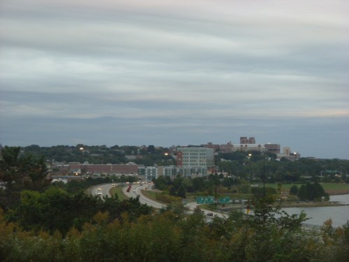 A view of a city skyline under cloudy skies, with a highway and greenery in the foreground.