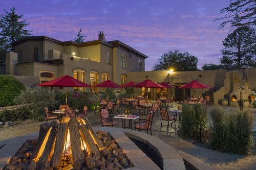 A cozy outdoor dining area with red umbrellas, a fire pit, and a beautiful sunset backdrop.