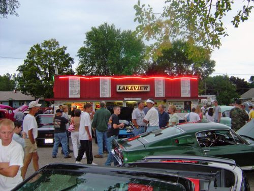 A lively crowd gathers outside Lakeview, a food stand, with classic cars parked nearby under a cloudy sky.