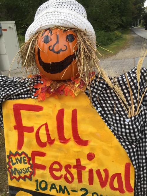 A cheerful scarecrow with a painted pumpkin head and straw hat stands next to a colorful sign for a Fall Festival.