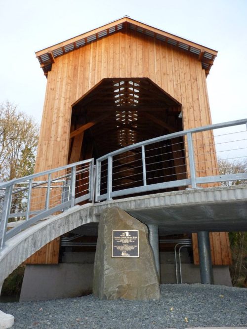 A wooden covered bridge with a stone base and plaque, set against a clear sky.