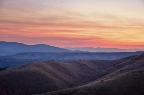 A serene landscape at sunset, featuring rolling hills and a colorful sky with soft pink and orange hues.