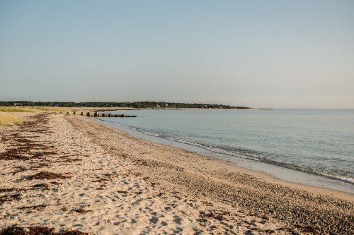 A serene beach scene with gentle waves, sandy shore, and a wooden pier extending into calm waters under a clear sky.