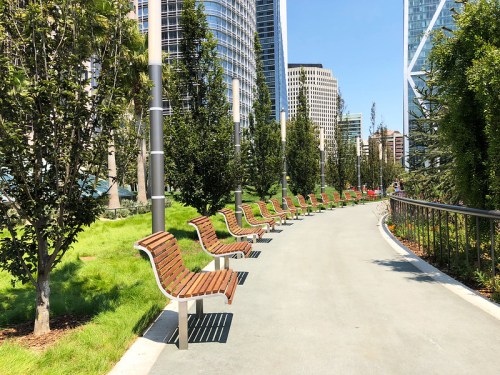 A sunny urban park pathway lined with modern wooden benches and greenery, surrounded by tall buildings.