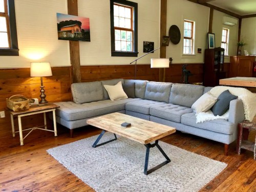 Cozy living room with a gray sectional sofa, wooden coffee table, and warm wooden accents. Natural light from windows.