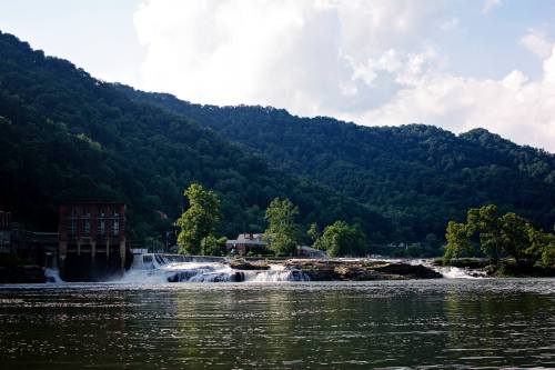 A serene river scene with a waterfall, lush green mountains, and a small building near the water's edge.