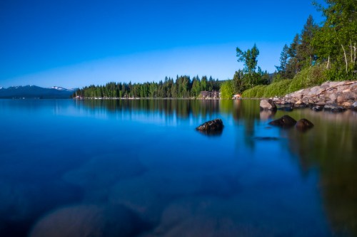 A serene lake scene with clear blue water, rocky shore, and lush green trees under a bright blue sky.