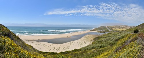 A panoramic view of a beach with gentle waves, sandy shore, and lush green hills under a clear blue sky.