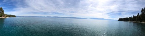 Panoramic view of a calm lake surrounded by trees and distant mountains under a clear blue sky.