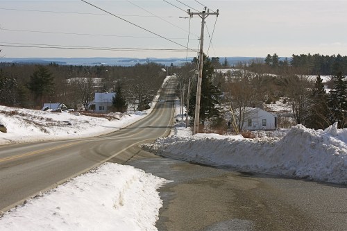 A snowy rural road stretches into the distance, lined with trees and houses under a clear sky.