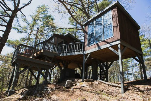 A wooden treehouse on stilts surrounded by trees, featuring large windows and a balcony.