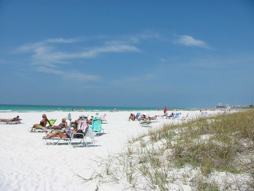 A sunny beach scene with people lounging on chairs, white sand, and turquoise water under a clear blue sky.
