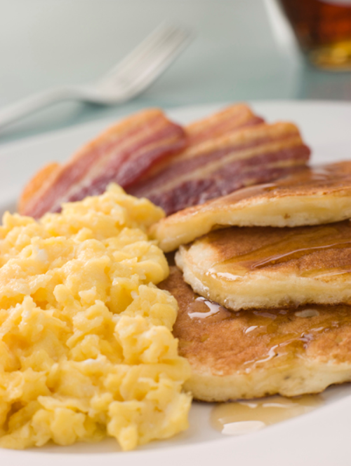 A plate of pancakes drizzled with syrup, scrambled eggs, and crispy bacon strips.