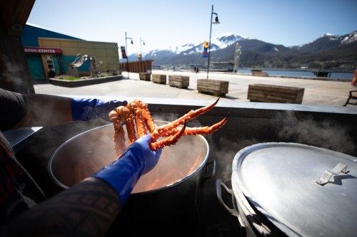 A person in blue gloves holds crab legs over a steaming pot, with a scenic waterfront and mountains in the background.