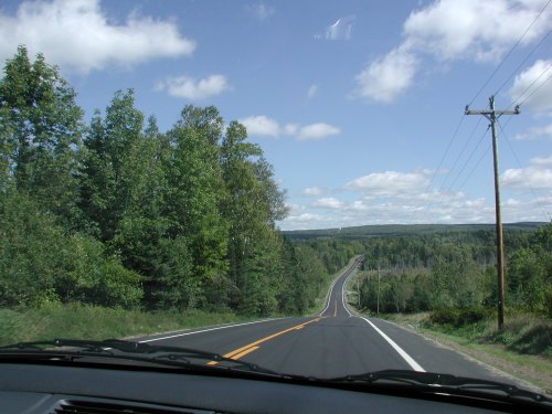 A winding road stretches through lush green trees under a partly cloudy sky.