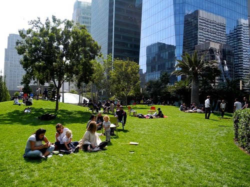 People relax on a green lawn in a city park, surrounded by tall buildings and palm trees on a sunny day.