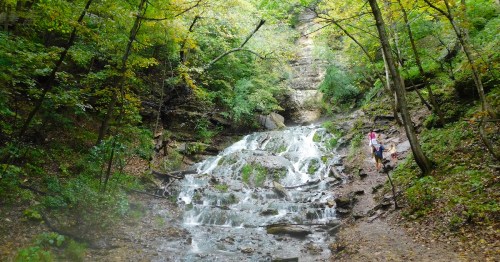 A serene waterfall cascades down rocky terrain, surrounded by lush green trees and a person walking nearby.