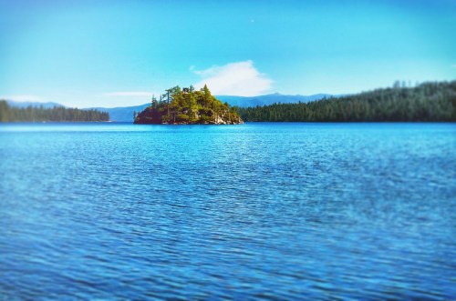 A serene lake scene featuring a small island with trees, surrounded by calm blue waters and distant mountains.