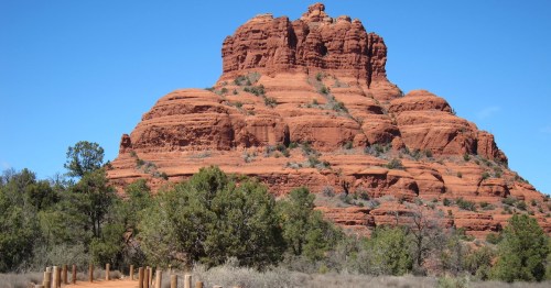 Red rock formation rises against a clear blue sky, surrounded by green shrubs and trees in a desert landscape.