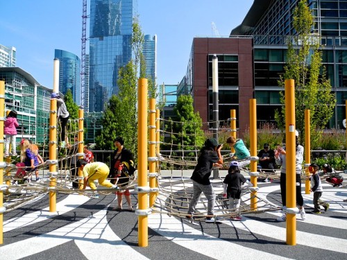 Children play on a climbing structure in a modern urban park, surrounded by tall buildings and greenery.