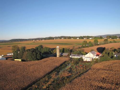 Aerial view of a rural landscape with fields, a farm, and a silo under a clear blue sky.