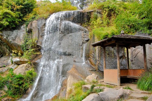 A serene waterfall cascades over rocky cliffs, beside a small wooden shelter surrounded by lush greenery.