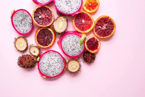 Colorful arrangement of dragon fruit, blood oranges, and rambutans on a pink background.