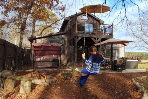 A person swings on a rope swing in a backyard with a house, patio, and fire pit surrounded by trees.