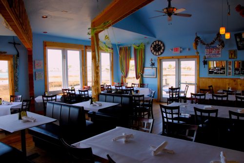 Brightly lit restaurant interior with blue walls, wooden beams, and tables set for dining, overlooking a scenic view.