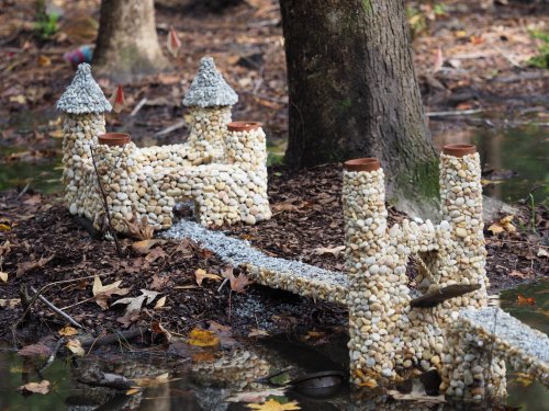 A small castle made of pebbles and stones, with towers and a bridge, situated in a wooded area by a pond.