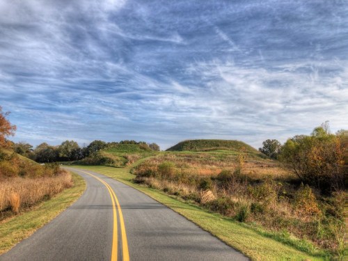 A winding road leads through grassy fields under a blue sky with wispy clouds, with a hill in the background.