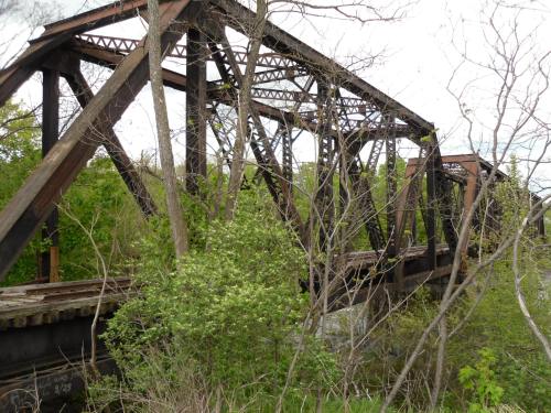 Rusty metal train bridge surrounded by greenery and trees, with a cloudy sky in the background.