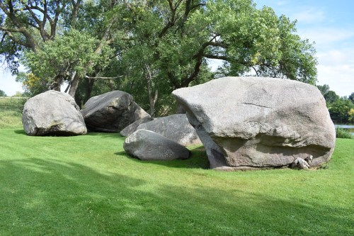Large boulders scattered on a grassy area, surrounded by trees and a clear blue sky.